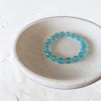 A beaded stretch bracelet made of translucent light blue beads separated by small gold spacer rings, displayed inside a shallow round ceramic dish on a textured off-white surface.