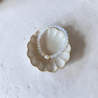 A beaded stretch bracelet with translucent white beads and a small gold accent, displayed inside a scalloped ceramic dish on a textured white surface.