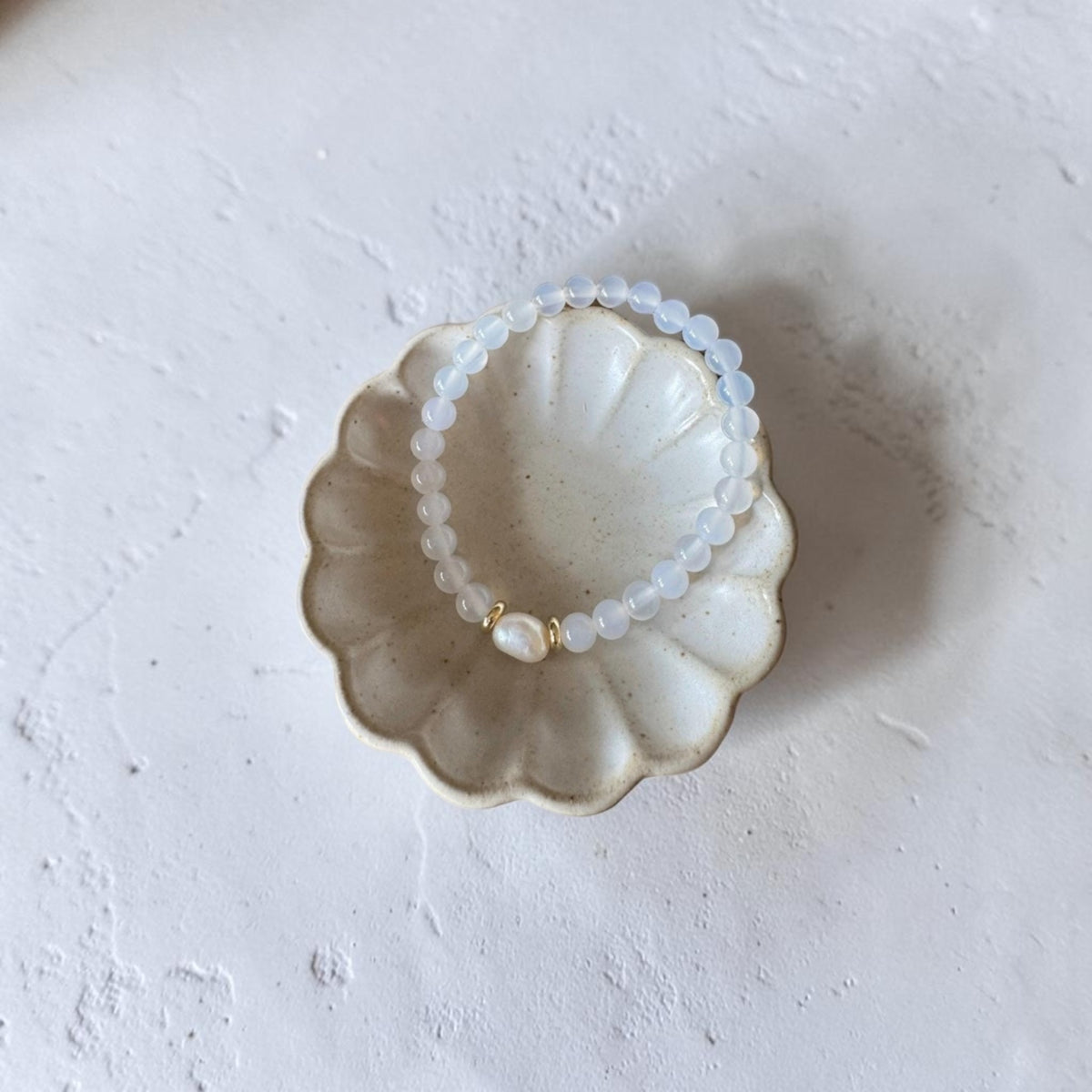 A beaded stretch bracelet with translucent white beads and a small gold accent, displayed inside a scalloped ceramic dish on a textured white surface.
