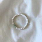 A beaded stretch bracelet featuring small white round beads and a single light green rectangular accent bead, displayed on a soft white fabric background.