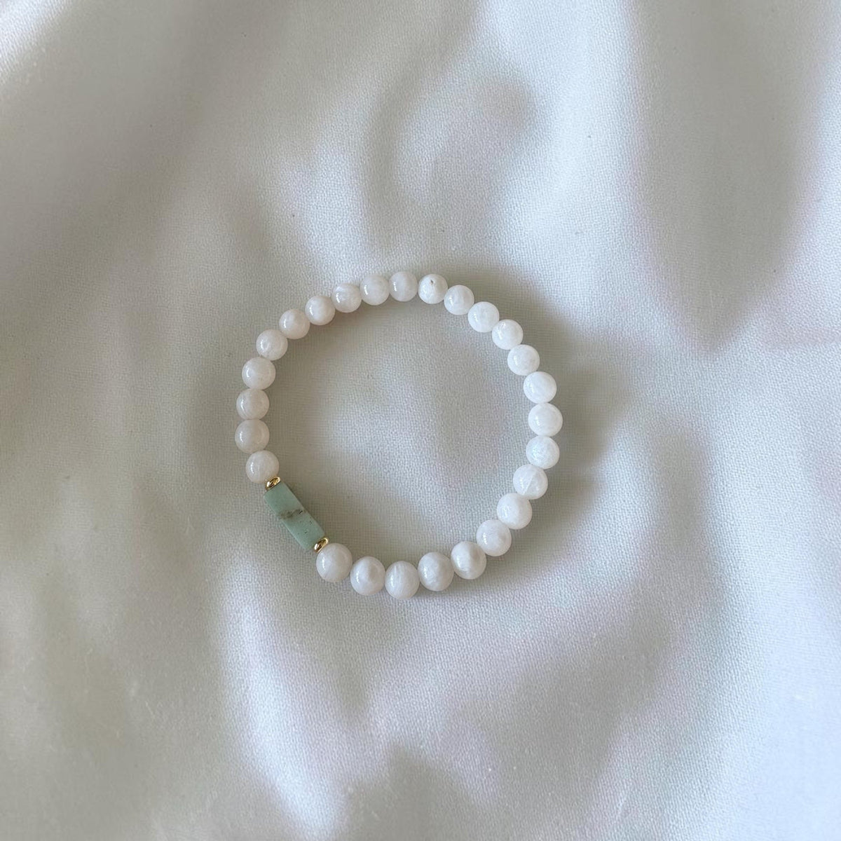 A beaded stretch bracelet featuring small white round beads and a single light green rectangular accent bead, displayed on a soft white fabric background.