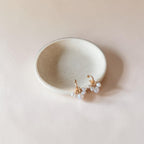 A pair of gold-toned dangle earrings with small white pearl-like beads, displayed on a small round ceramic dish against a warm beige background.