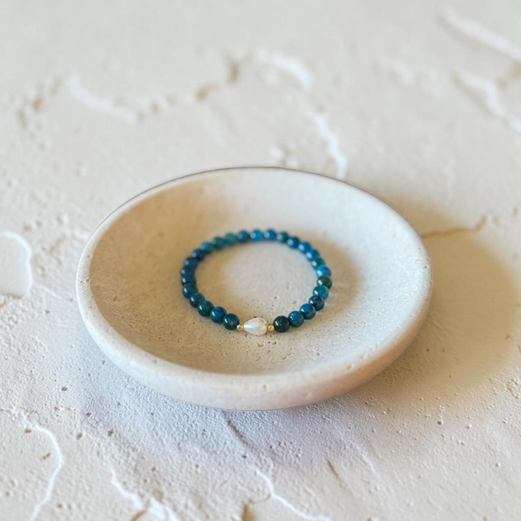 A beaded stretch bracelet with dark teal-blue beads and a small white pearl accent, displayed on a small round ceramic dish over a textured beige surface.
