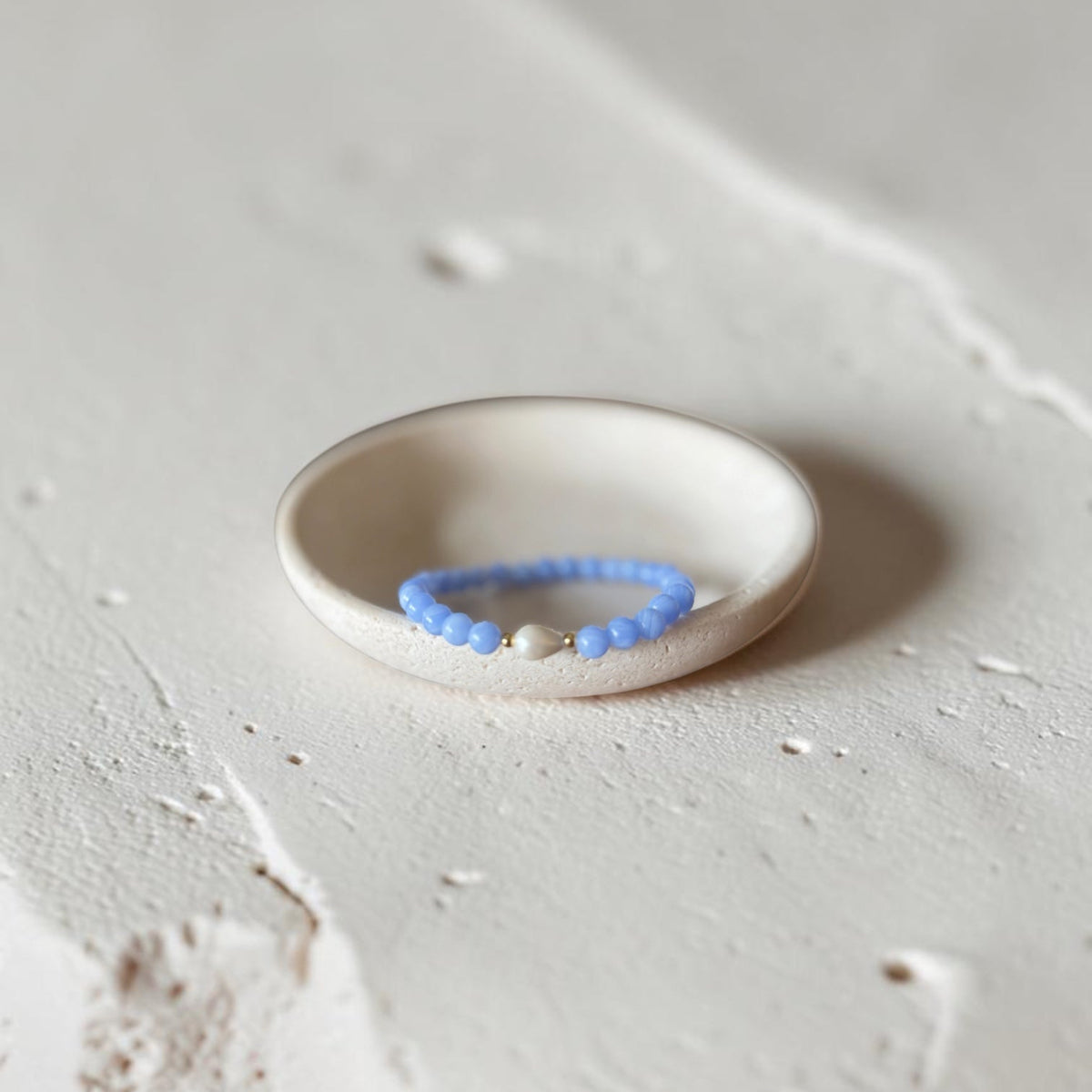 A delicate beaded bracelet with light blue beads and a small white stone, displayed inside a small white ceramic dish on a textured light gray surface.