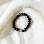 A beaded stretch bracelet with glossy black round beads and a small white spacer bead with black speckles, displayed on a soft white fabric background.