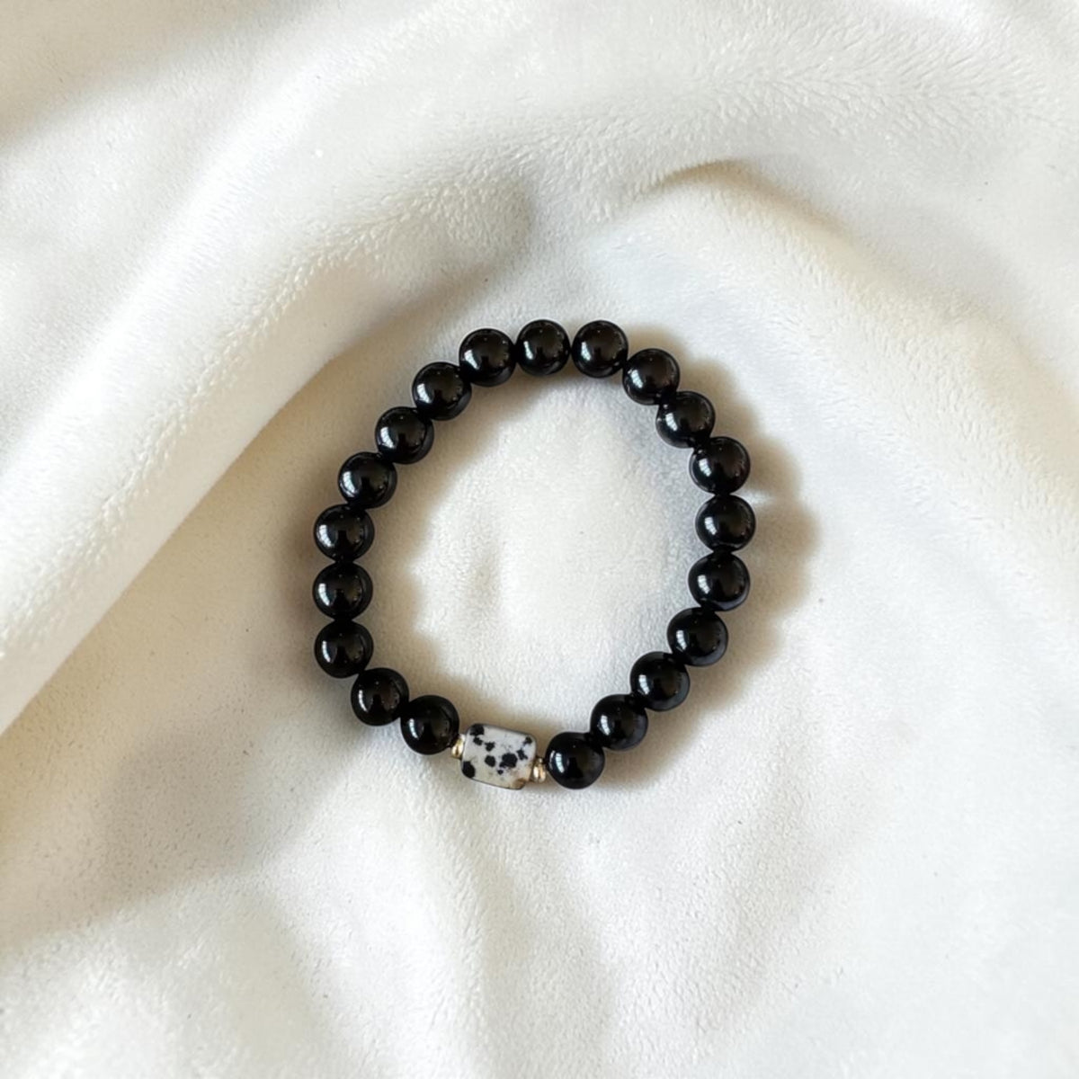 A beaded stretch bracelet with glossy black round beads and a small white spacer bead with black speckles, displayed on a soft white fabric background.