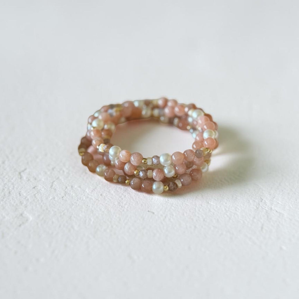 A multi-strand beaded ring featuring pink, peach, and iridescent white beads, photographed on a textured off-white surface.