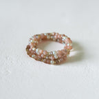 A multi-strand beaded ring featuring pink, peach, and iridescent white beads, photographed on a textured off-white surface.