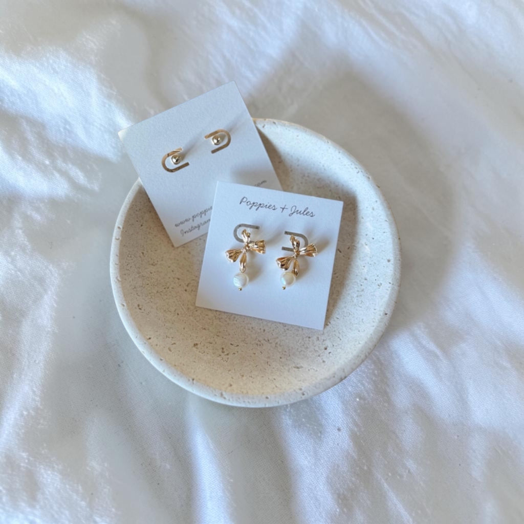 Two pairs of gold-toned dangle earrings displayed on small white cards, resting in a round speckled ceramic dish on a white fabric background.