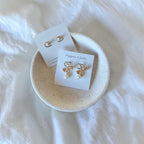 Two pairs of gold-toned dangle earrings displayed on small white cards, resting in a round speckled ceramic dish on a white fabric background.