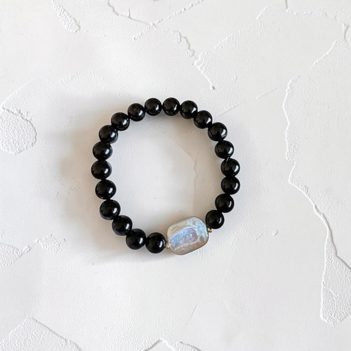 A beaded stretch bracelet featuring glossy black round beads and a central irregular white stone with iridescent blue-green highlights, displayed on a textured off-white surface.