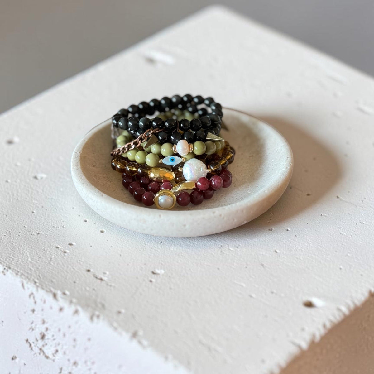 A stack of beaded bracelets in black, green, red, and gold tones displayed on a small round white dish, set on a textured white block against a neutral gray background.