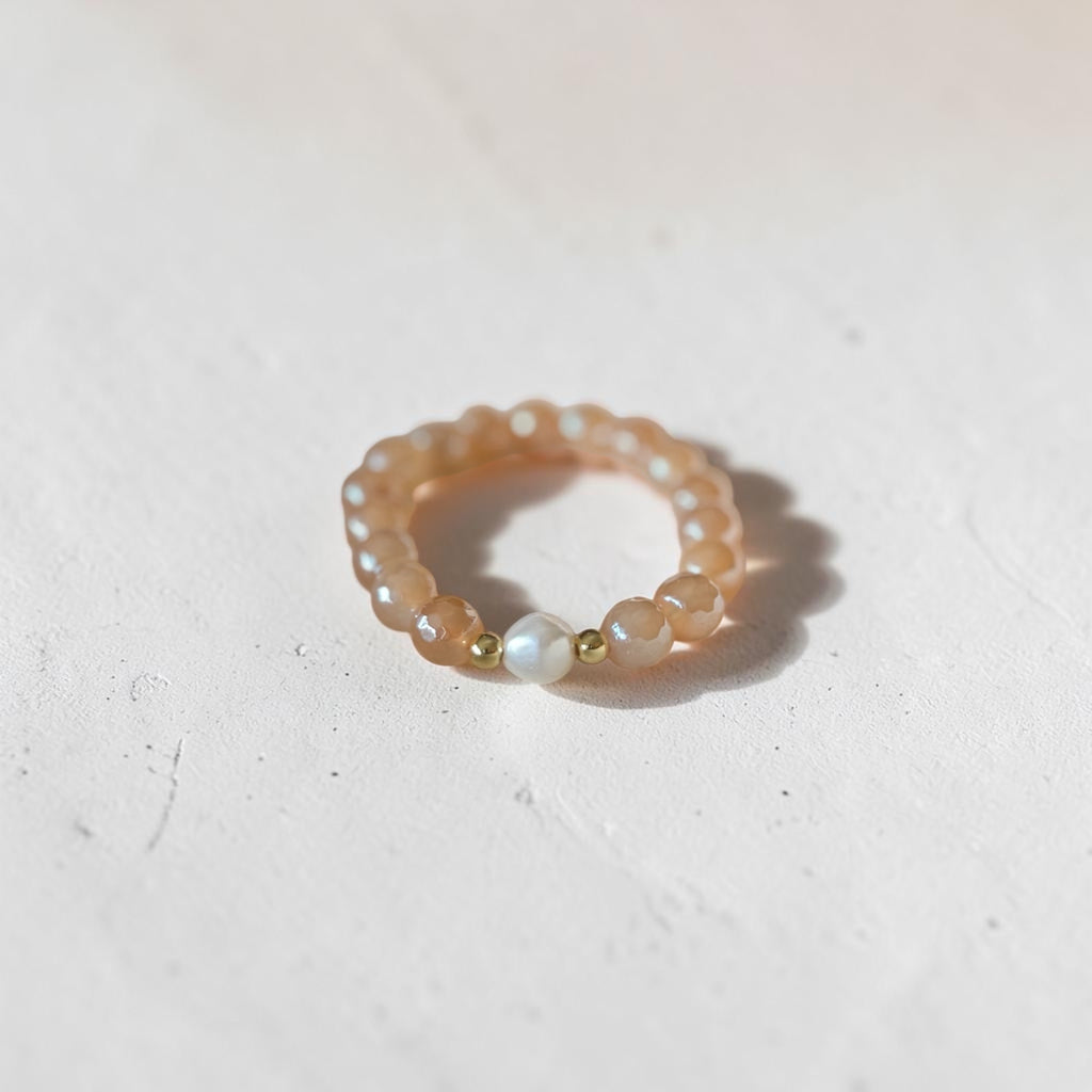 A beaded stretch bracelet featuring peachy-orange translucent beads, small white pearl-like beads, and two gold spacer beads, resting on a textured off-white surface.