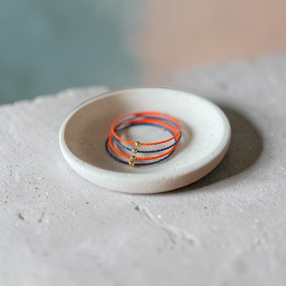 4 delicate wire bracelets in orange, navy accented with small gold beads, displayed on a small round ceramic dish.