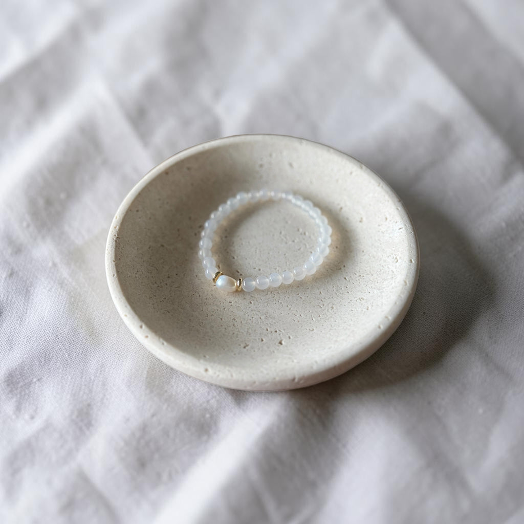 A small round ceramic dish holding a delicate white beaded bracelet with a tiny gold accent, set on a textured white fabric background.