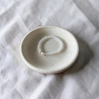 A small round ceramic dish holding a delicate white beaded bracelet with a tiny gold accent, set on a textured white fabric background.