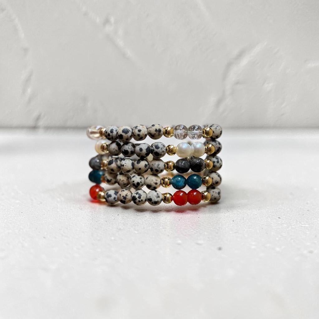 A stack of beaded stretch bracelets displayed on a white surface against a textured white wall. The bracelets feature speckled gray and black beads, white beads, gold spacer beads, and small accent beads in red, teal blue, and white.