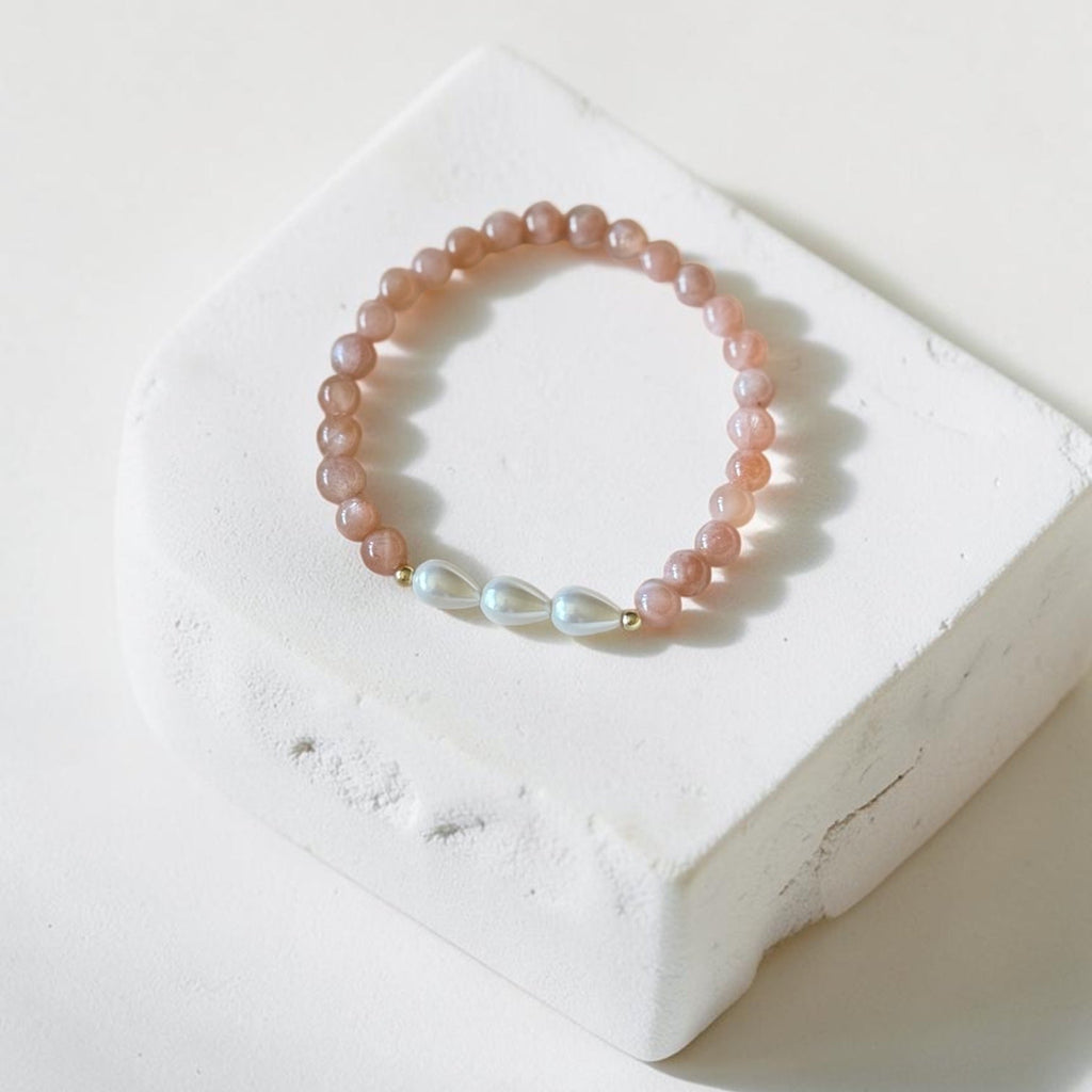 A beaded stretch bracelet in soft dusty pink with three iridescent white pearl-like beads and a small gold accent, displayed on a white textured block against a warm off-white background.