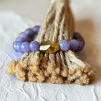 A beaded bracelet with smooth lavender-purple round beads and a faceted metallic gold accent bead, displayed on a textured beige woven mat against a light neutral background.