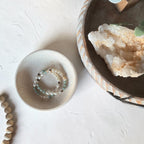 Two beaded stretch bracelets with light blue, white, and brown beads displayed in a small speckled ceramic dish, styled on a textured white surface with boho props including a wooden tray, crystal, and beads.