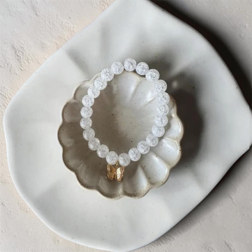 A beaded stretch bracelet made of clear faceted round beads with a small gold-toned connector, displayed on a scalloped white ceramic dish against a light textured background.