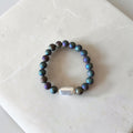 A beaded stretch bracelet featuring iridescent dark teal and purple pearls with a central white baroque bead, displayed on a light marble surface.