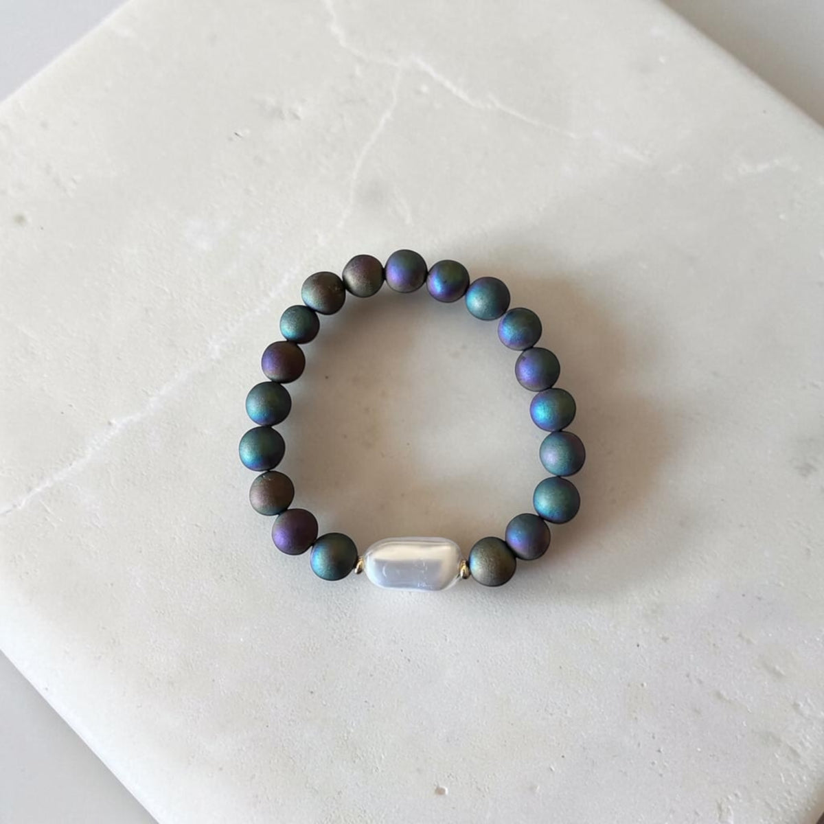 A beaded stretch bracelet featuring iridescent dark teal and purple pearls with a central white baroque bead, displayed on a light marble surface.