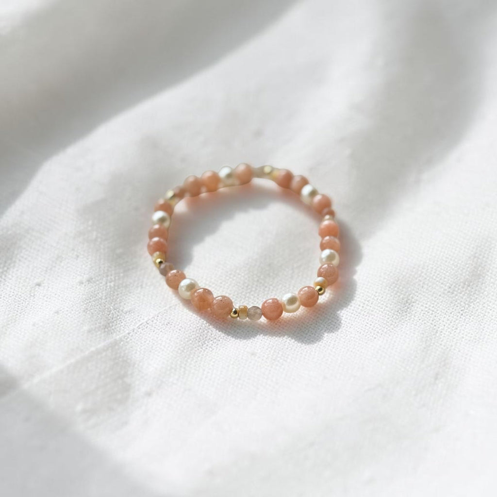 A delicate beaded ring with peach-pink and white beads, accented with small gold spacer beads, resting on a softly draped white fabric background.