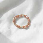 A delicate beaded ring with peach-pink and white beads, accented with small gold spacer beads, resting on a softly draped white fabric background.