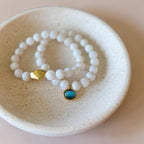 Two beaded stretch bracelets with white beads and gold-toned accents, displayed in a round beige ceramic dish. One bracelet features a faceted gold bead, and the other has a round bezel-set turquoise stone.