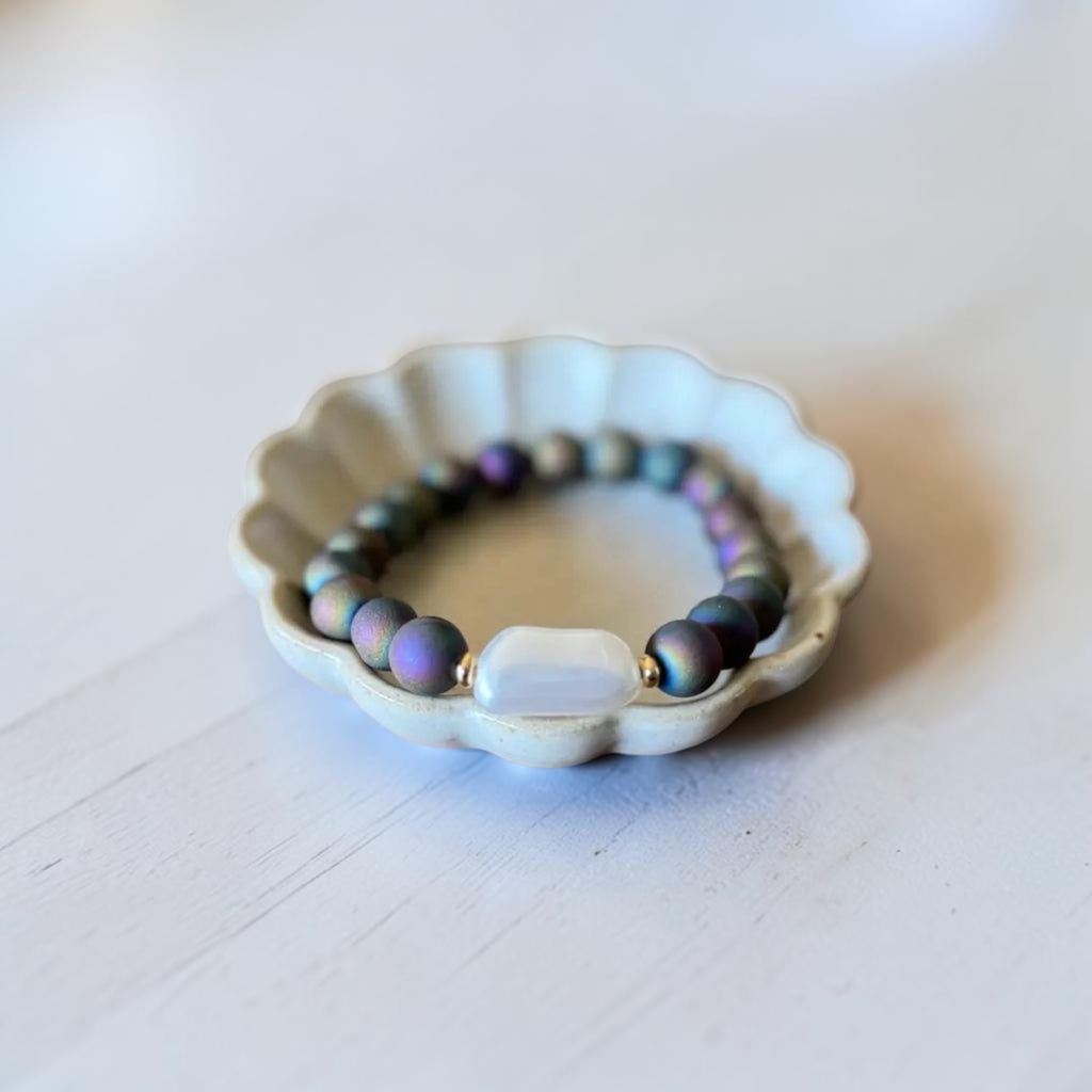A beaded stretch bracelet with iridescent dark beads and a central white oval stone, displayed on a small white scalloped dish over a light surface.