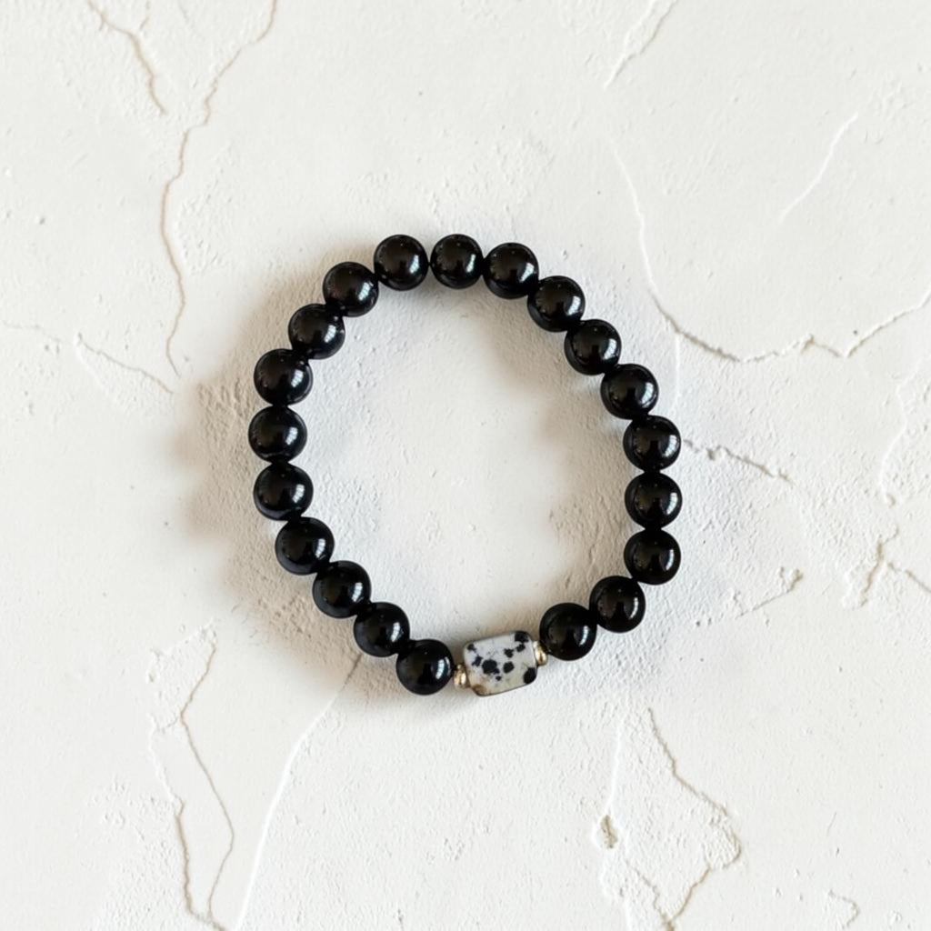 A beaded stretch bracelet featuring glossy black round beads and a single white Dalmatian-patterned accent bead, displayed on a textured off-white surface.