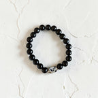 A beaded stretch bracelet featuring glossy black round beads and a single white Dalmatian-patterned accent bead, displayed on a textured off-white surface.