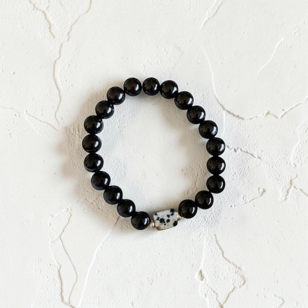 A beaded stretch bracelet featuring glossy black round beads and a single white Dalmatian-patterned accent bead, displayed on a textured off-white surface.