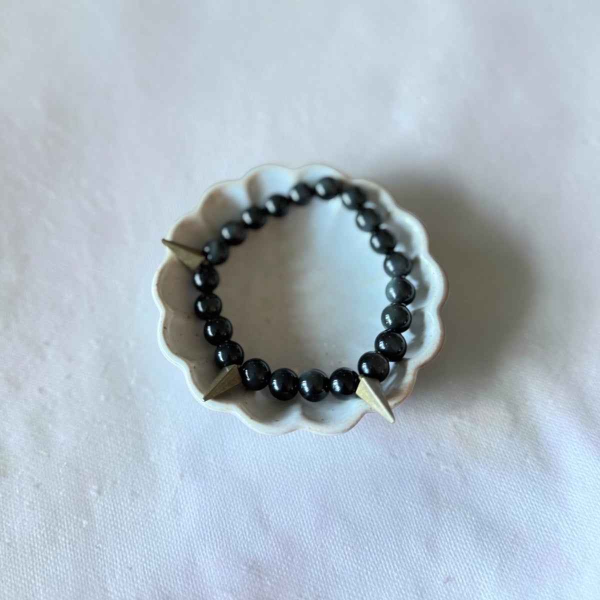 A beaded stretch bracelet with glossy black round beads and two small gold-toned metal spikes, displayed on a small white scalloped dish over a light fabric background.