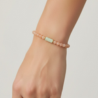 A close-up of a person's wrist wearing a delicate beaded bracelet. The bracelet consists of small, round dusty pink beads with a central, slightly larger pale mint-green rectangular bead.