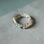 A delicate beaded ring with small white pearls and pale green beads, resting on a textured gray concrete surface.