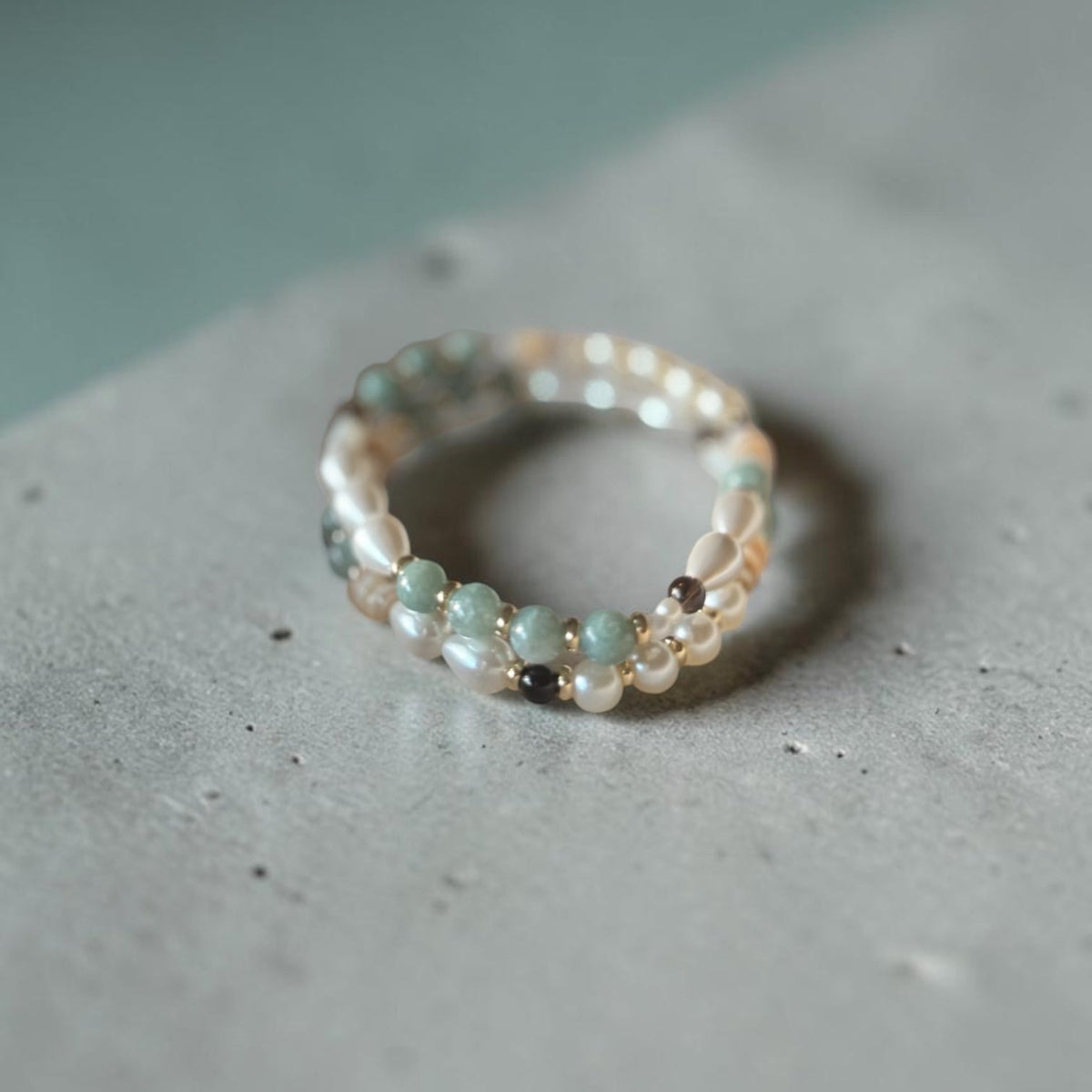 A delicate beaded ring with small white pearls and pale green beads, resting on a textured gray concrete surface.