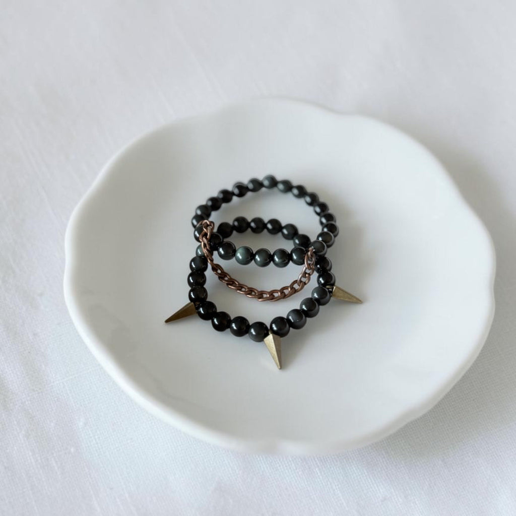 Two beaded stretch bracelets displayed on a small white ceramic dish. The bracelets feature black beads, dark gray beads, a bronze-toned chain, and gold-toned spike charms.