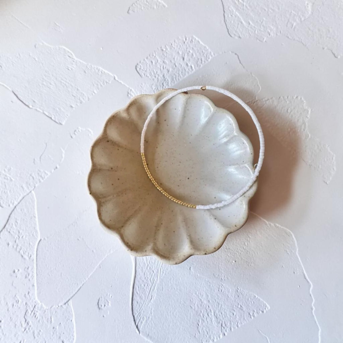 A pair of delicate beaded wire bracelet, white beads and cluster in gold beads, displayed inside a scalloped ceramic dish on a textured white surface.