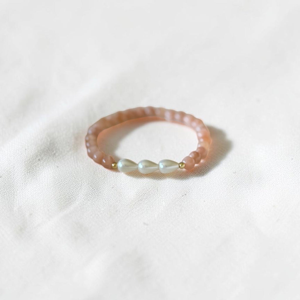 A delicate beaded ring featuring a peach-pink translucent band with three small white pearl-like beads separated by tiny gold spacer beads, resting on a soft white fabric background.
