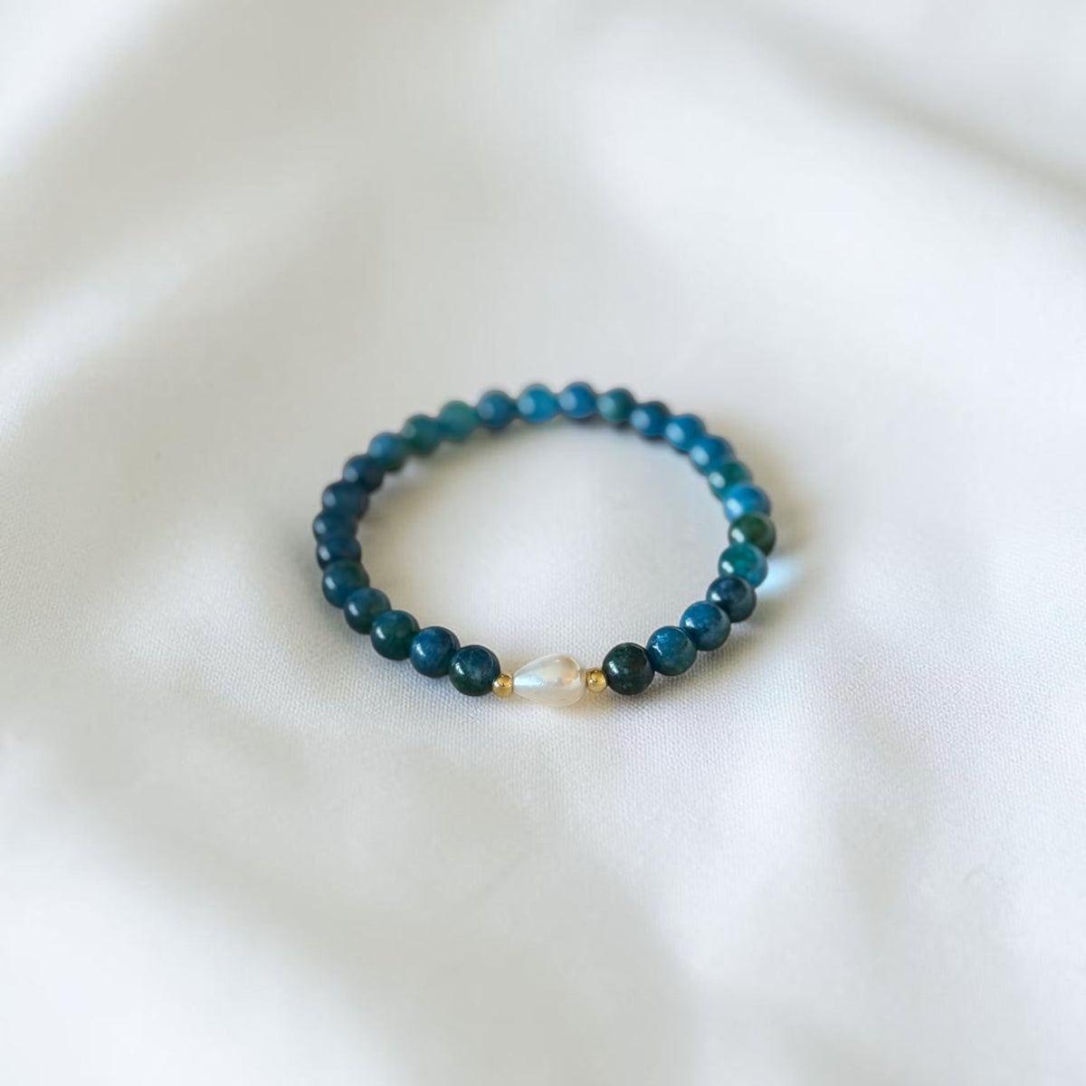 A beaded stretch bracelet with dark teal-blue beads and a small iridescent pearl accent, resting on a soft white fabric background.