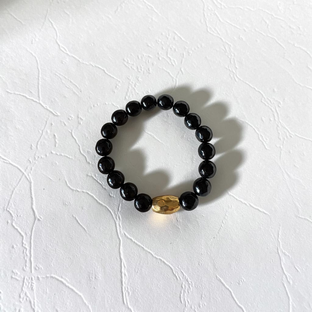 A beaded stretch bracelet featuring round black beads and a single faceted gold accent bead, displayed on a textured white surface.
