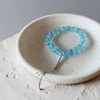 A light blue beaded bracelet with a silver-toned chain detail, displayed inside a small speckled ceramic dish on a textured light surface.