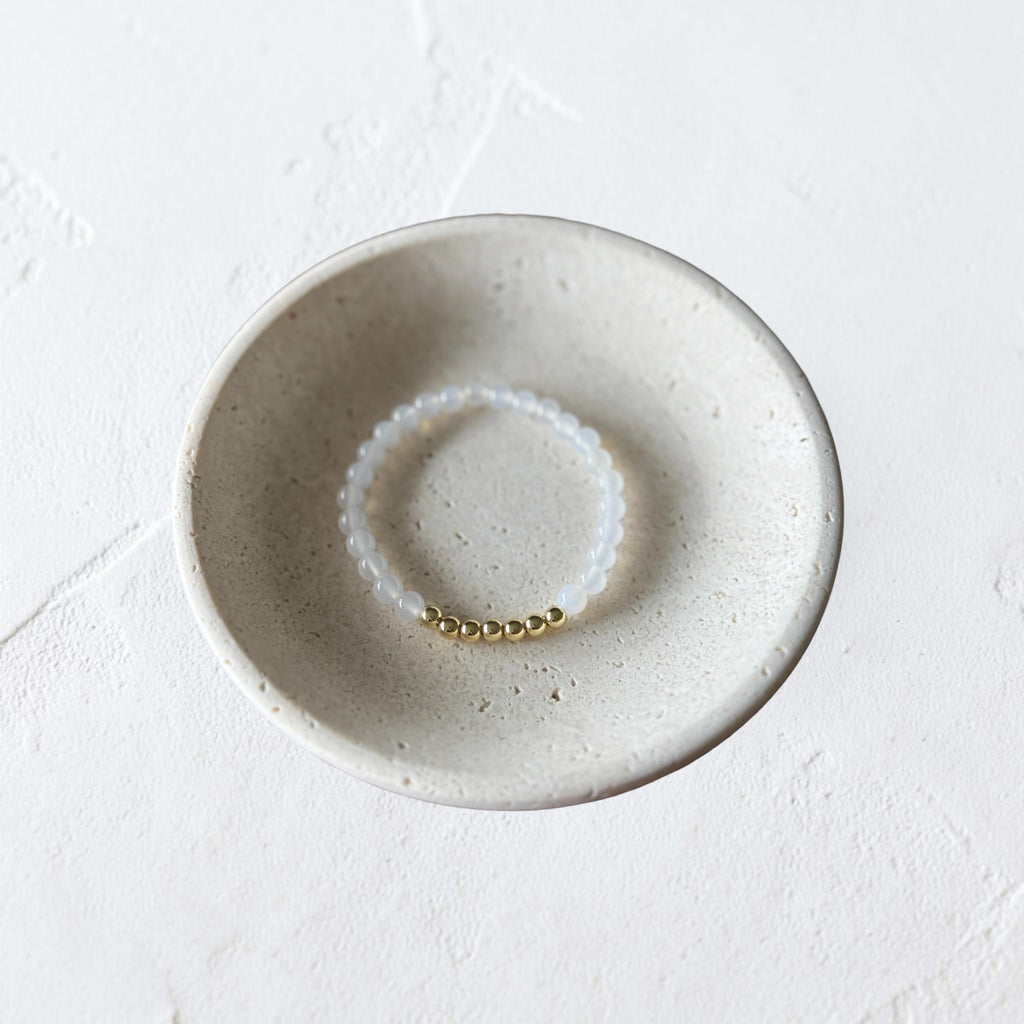 A delicate beaded bracelet with white translucent beads and a small gold bead accent, displayed inside a round, speckled ceramic dish on a textured white surface.