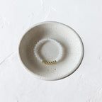 A delicate beaded bracelet with white translucent beads and a small gold bead accent, displayed inside a round, speckled ceramic dish on a textured white surface.