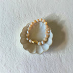 A beaded stretch bracelet with cream, white, with swirl beads and a small gold accent bead, displayed on a scalloped ceramic dish against a textured off-white background.