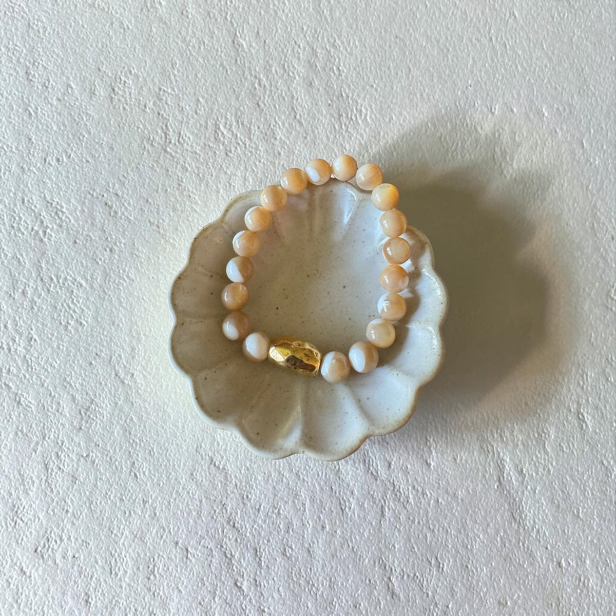 A beaded stretch bracelet with cream, white, with swirl beads and a small gold accent bead, displayed on a scalloped ceramic dish against a textured off-white background.