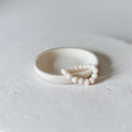 A minimalist cream-colored ceramic ring with a small beaded accent, photographed on a textured light gray surface.