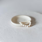A minimalist cream-colored ceramic ring with a small beaded accent, photographed on a textured light gray surface.